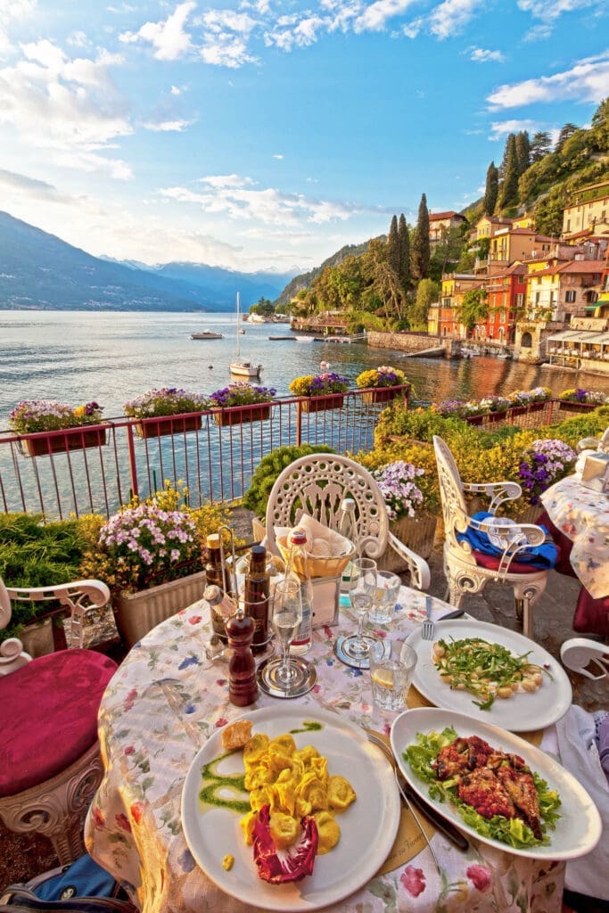 Outdoor dining overlooking Lake Como with colorful waterfront villas, mountains, and scenic Italian lakeside views.