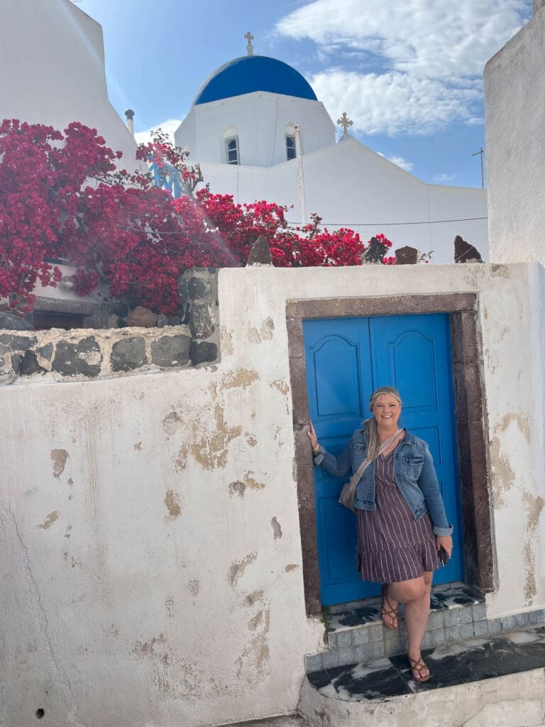 Stacy, founder of Epic Sands Travel, standing beside a blue door beneath a traditional blue-domed church in Santorini, Greece.